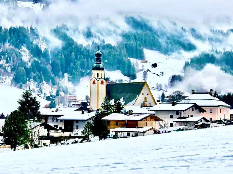 Die Kirche von Jochberg mit dem verschneiten Dorf und dem bewaldeten Berghang im Nebel